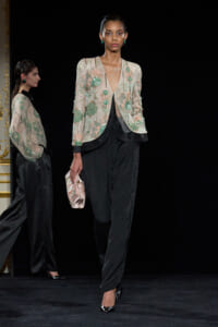 Model walking a runway in a sheer embellished jacket with green beads over a black outfit, holding a pale clutch with black heels.