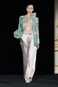 Model on a runway wearing a mint blazer with a sheer pastel blouse, pale pink satin wide-leg pants, and a matching green clutch.