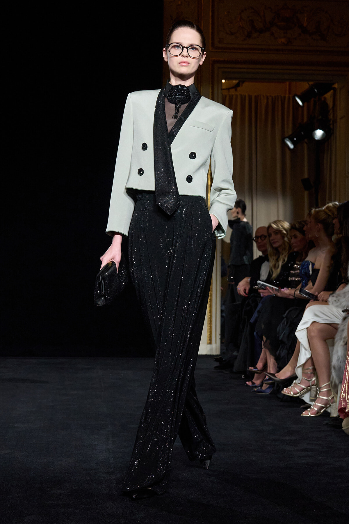 Model walks the runway in a pale gray cropped double-breasted blazer paired with glittery black wide-leg pants and a black scarf, wearing glasses and holding a clutch.