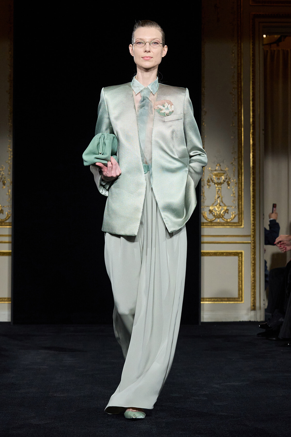 Model on a fashion runway wearing a mint-green satin suit with an oversized blazer, matching blouse and wide-leg trousers, holding a green clutch.