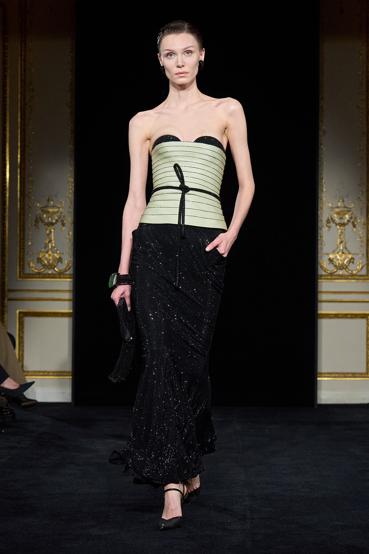 Model walks a runway wearing a strapless mint corset with black trim and a sparkling black gown, holding a clutch.