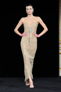 Model walks a runway wearing a fitted, sheer-beaded nude gown with ornate silver embroidery and a green pendant; she has turquoise-green earrings and beige heels, hands on hips.