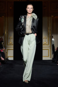 Model walks the runway in mint satin wide-leg pants, sheer blouse, and a black embroidered blazer with a large bow at the neck.