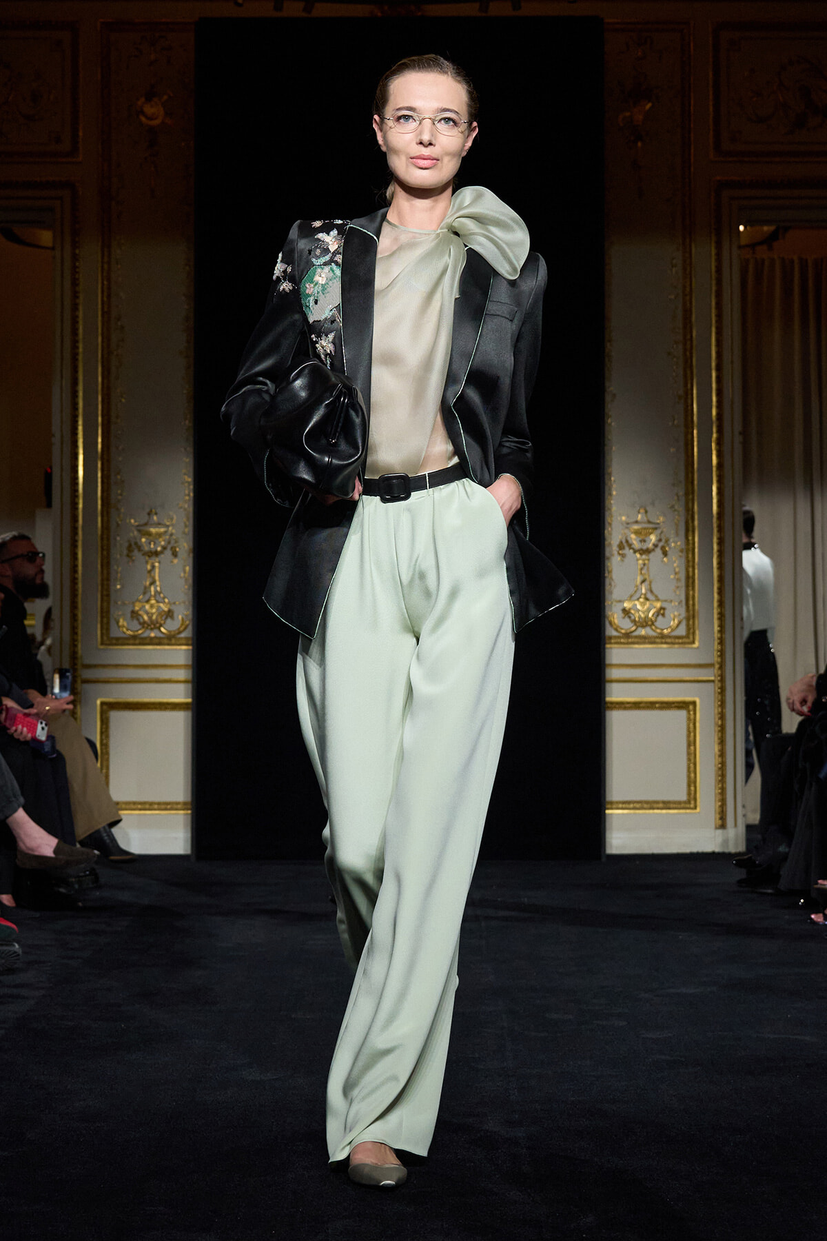 Model walks the runway in mint satin wide-leg pants, sheer blouse, and a black embroidered blazer with a large bow at the neck.