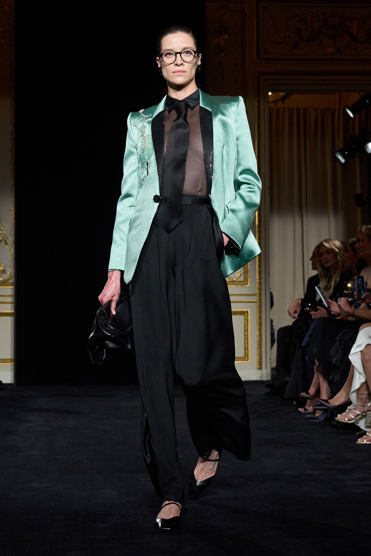 Model on a runway wearing a mint-green blazer over a sheer black blouse and black wide-legged pants, carrying a black clutch.