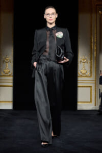 Model on a runway wearing a tailored black suit with a sheer blouse, round glasses, and a decorative brooch, holding a black clutch.