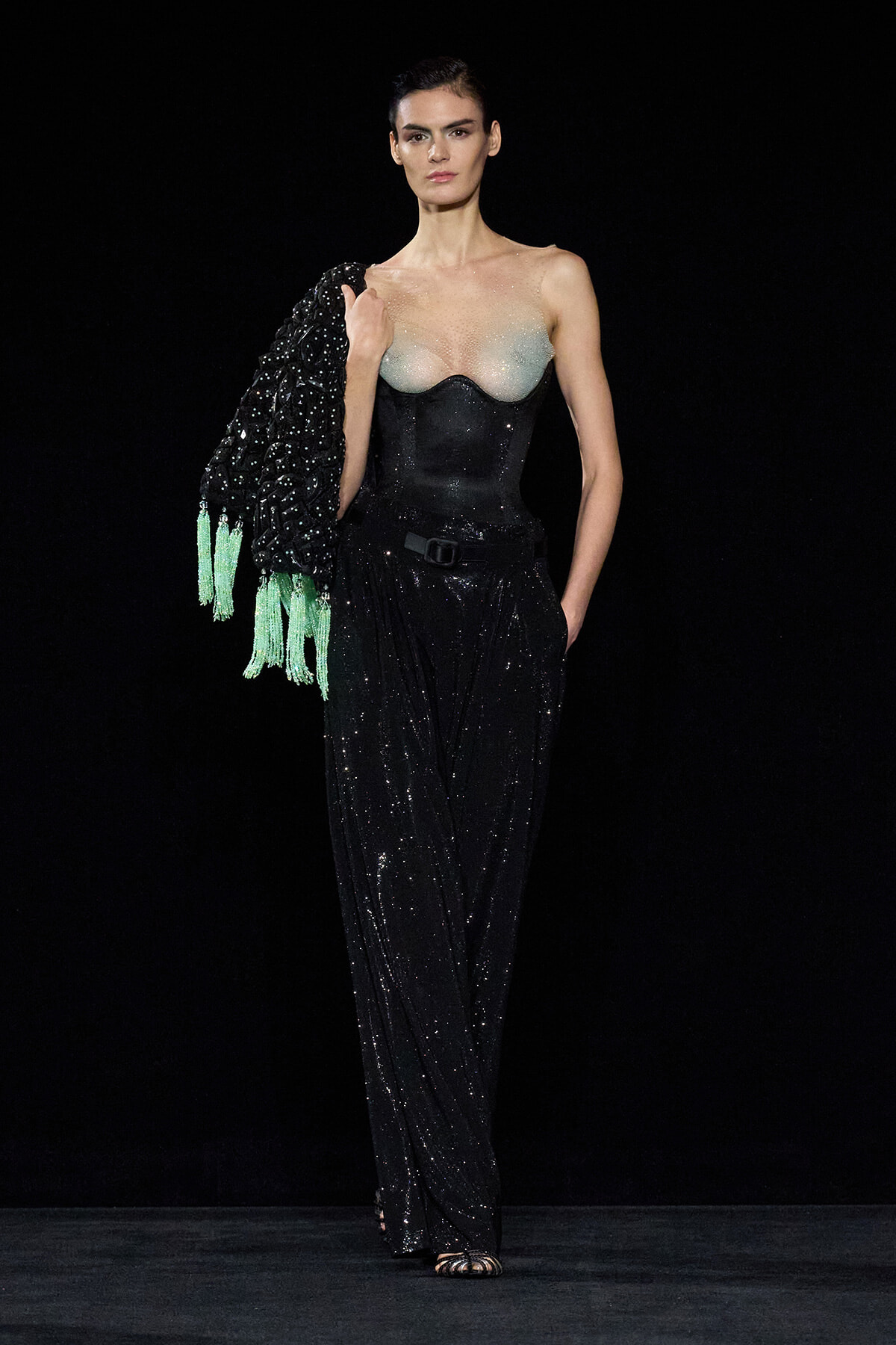 Model on a dark runway wearing a black sequined jumpsuit with a pale sheer bustier, holding a black beaded jacket with green tassels over her shoulder.