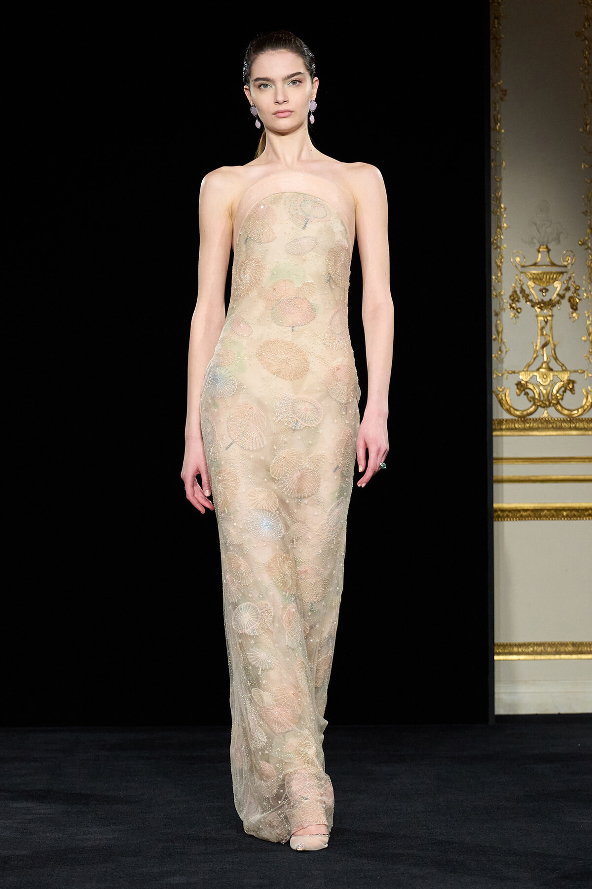 Model on a runway wearing a strapless, floor-length beige gown with colorful floral embroidery and delicate sheer overlay.