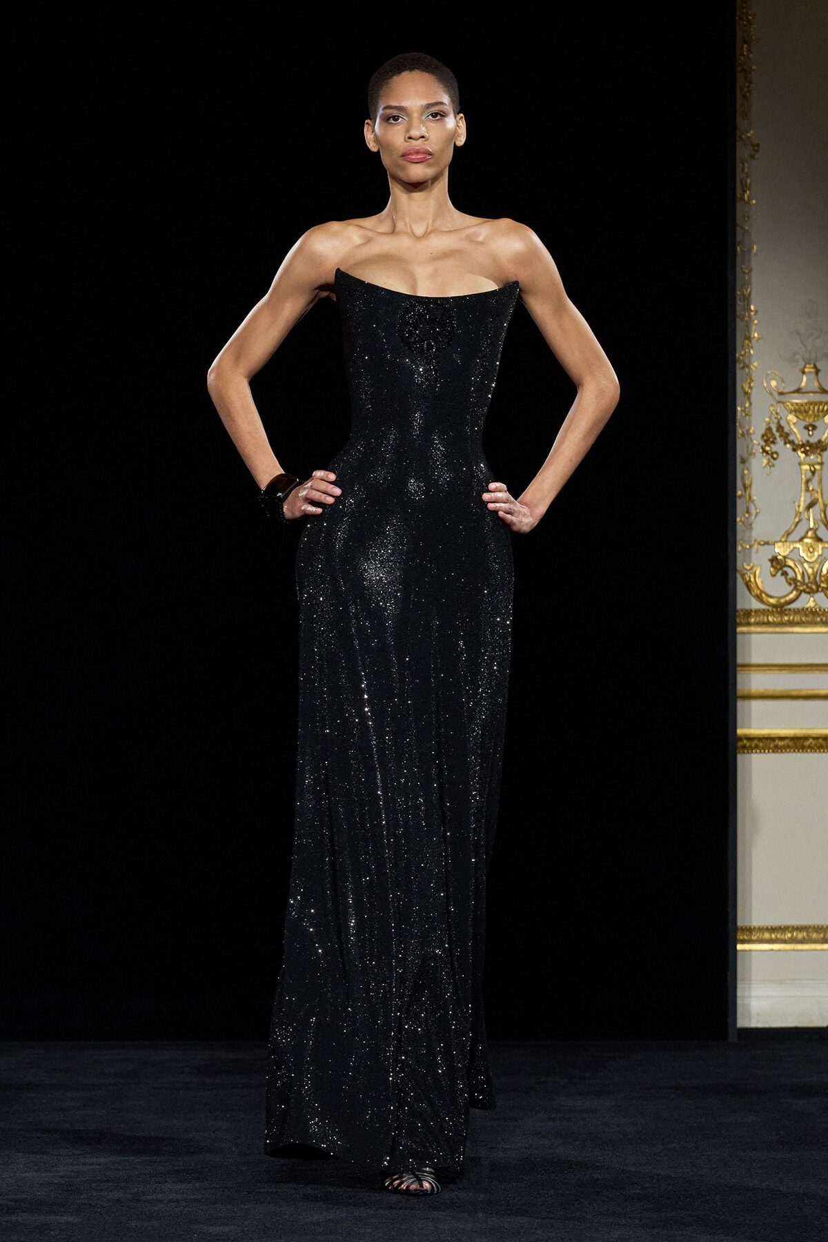 Model on a runway wearing a floor-length, black sequined gown with a structured, sweetheart strapless neckline and hands on hips.