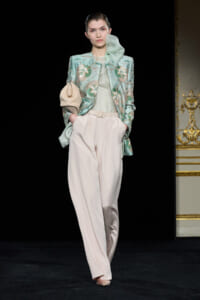 Model walks the runway in a mint-green embroidered jacket over a pale green top, paired with high-waisted cream trousers and a beige clutch weaved under her arm.