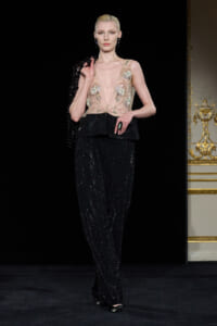 Model on a runway wearing a sheer nude top with floral embroidery and a layered, glittery black pant set.