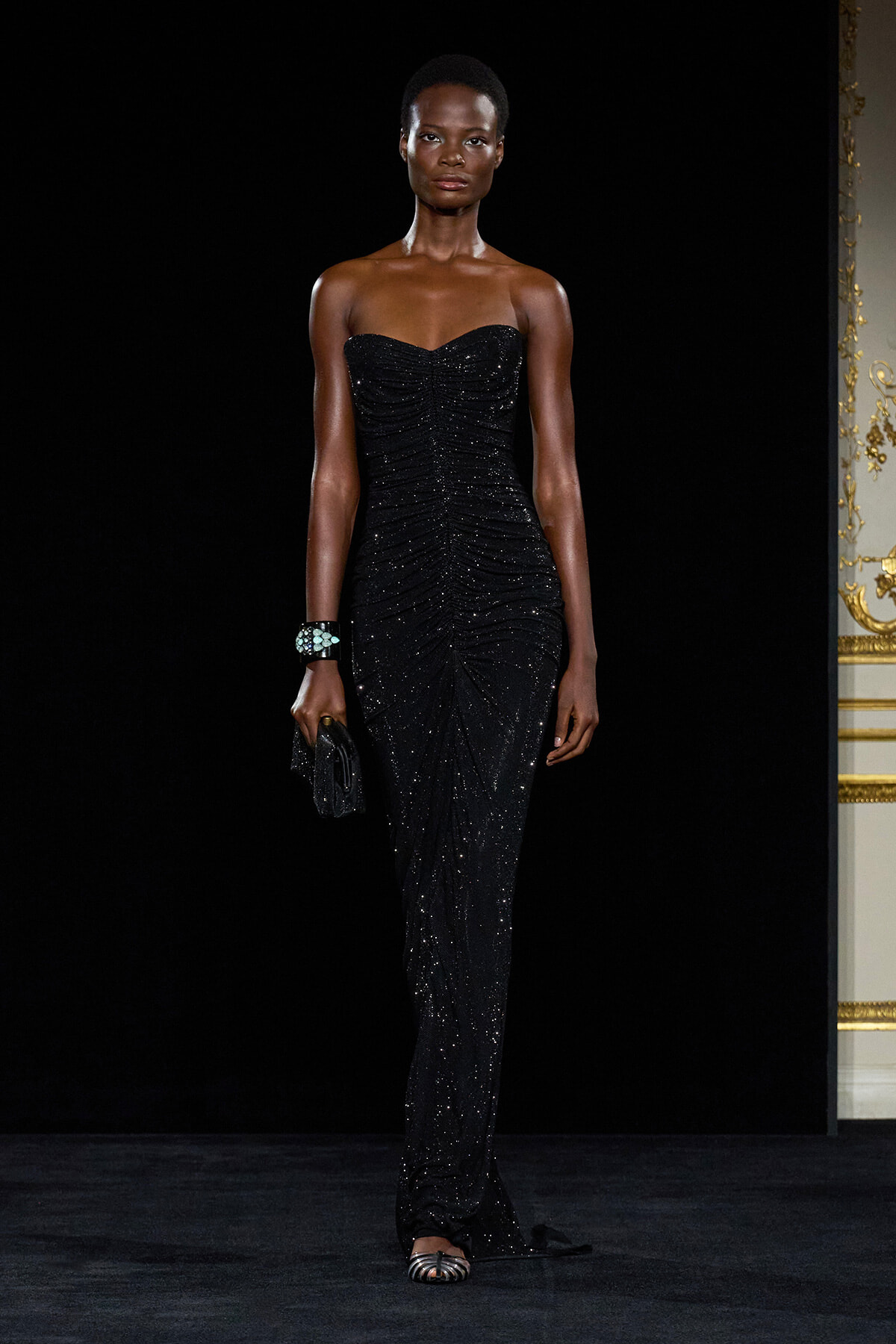 Model walks a runway in a strapless black, sequined gown with a ruched bodice, holding a small clutch and wearing a turquoise-stone bracelet.