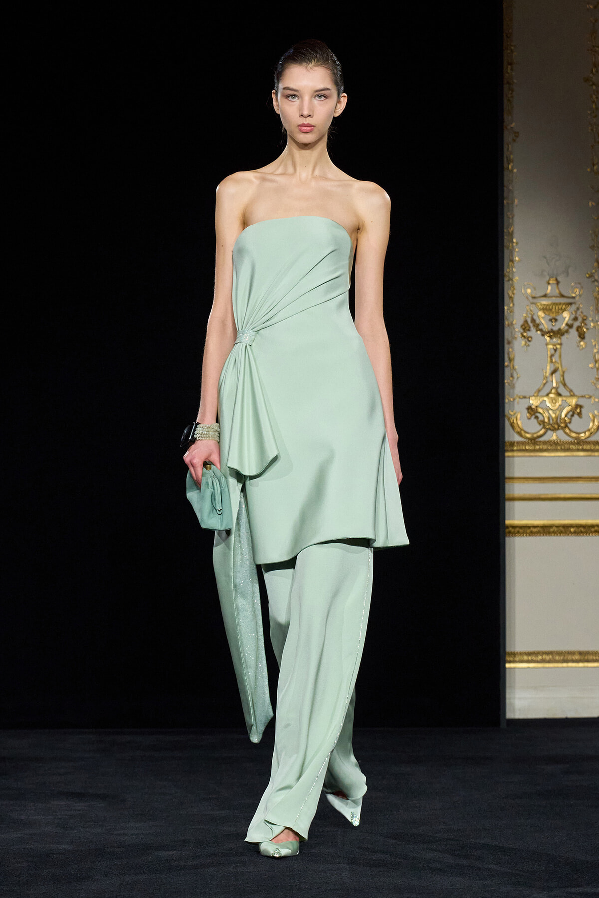 Model on a runway wearing a strapless mint-green draped ensemble with a side gathered detail and matching pants, holding a mint clutch.
