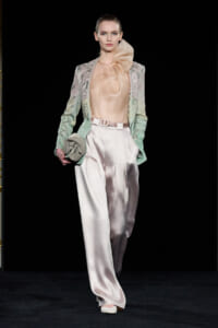 Model on a fashion runway wearing a pale peach satin blouse with a large bow, a mint lace blazer, and ivory wide-leg satin trousers, holding a small clutch.
