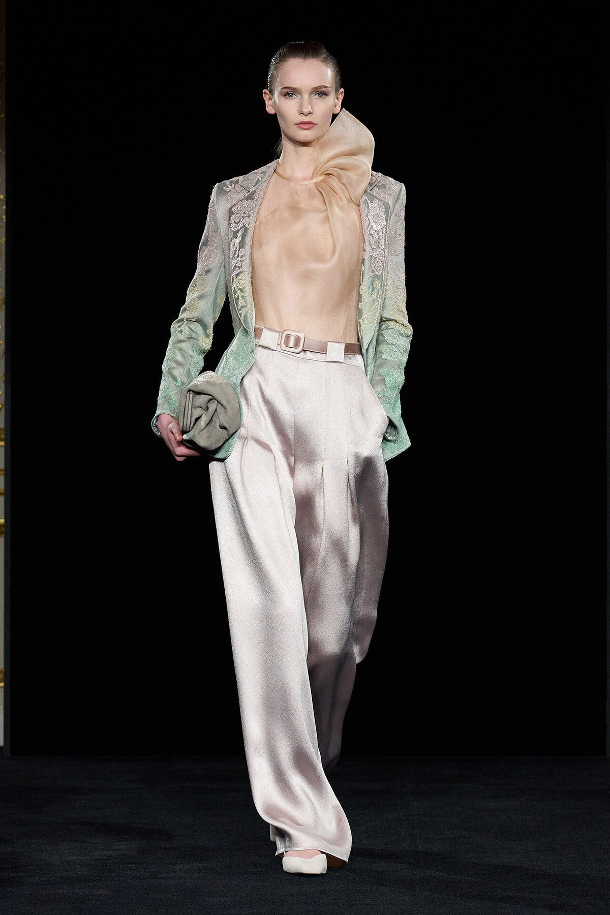 Model on a fashion runway wearing a pale peach satin blouse with a large bow, a mint lace blazer, and ivory wide-leg satin trousers, holding a small clutch.