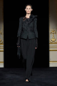 Model walking a runway in an all-black ensemble: textured blazer with a satin bow, sheer panel at the neckline, and wide-legged trousers, complemented by glittery gloves.