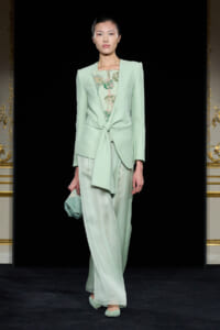 Model in mint green tailored suit with a front knot, sheer patterned top, and wide-leg pants on a runway, carrying a matching clutch.