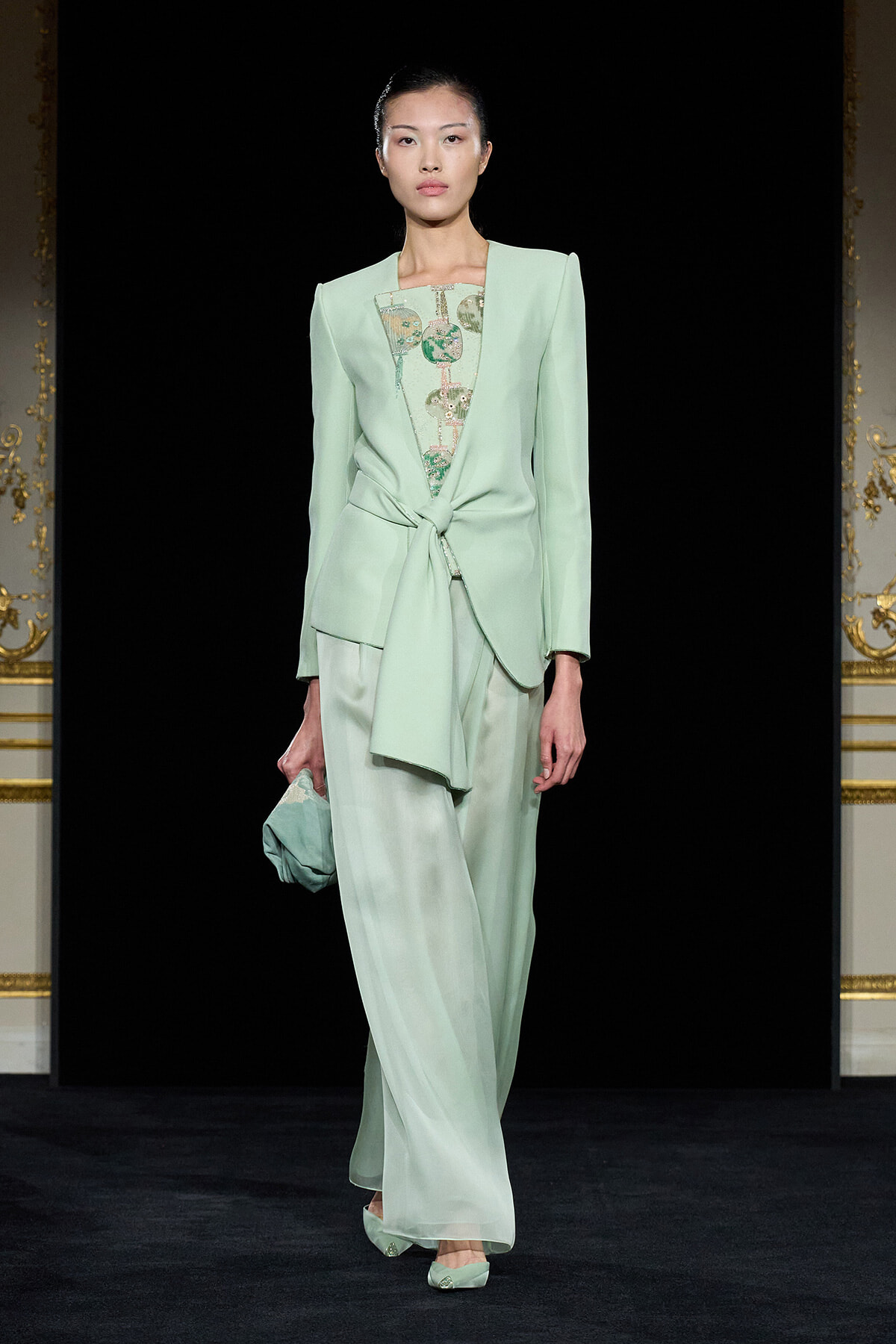 Model in mint green tailored suit with a front knot, sheer patterned top, and wide-leg pants on a runway, carrying a matching clutch.