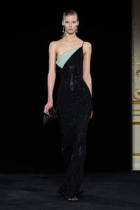Model on a runway wearing a black sequined, one-shoulder gown with a mint-green panel, holding a small black clutch.