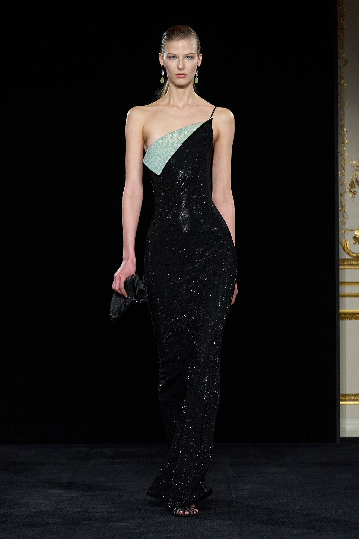 Model on a runway wearing a black sequined, one-shoulder gown with a mint-green panel, holding a small black clutch.