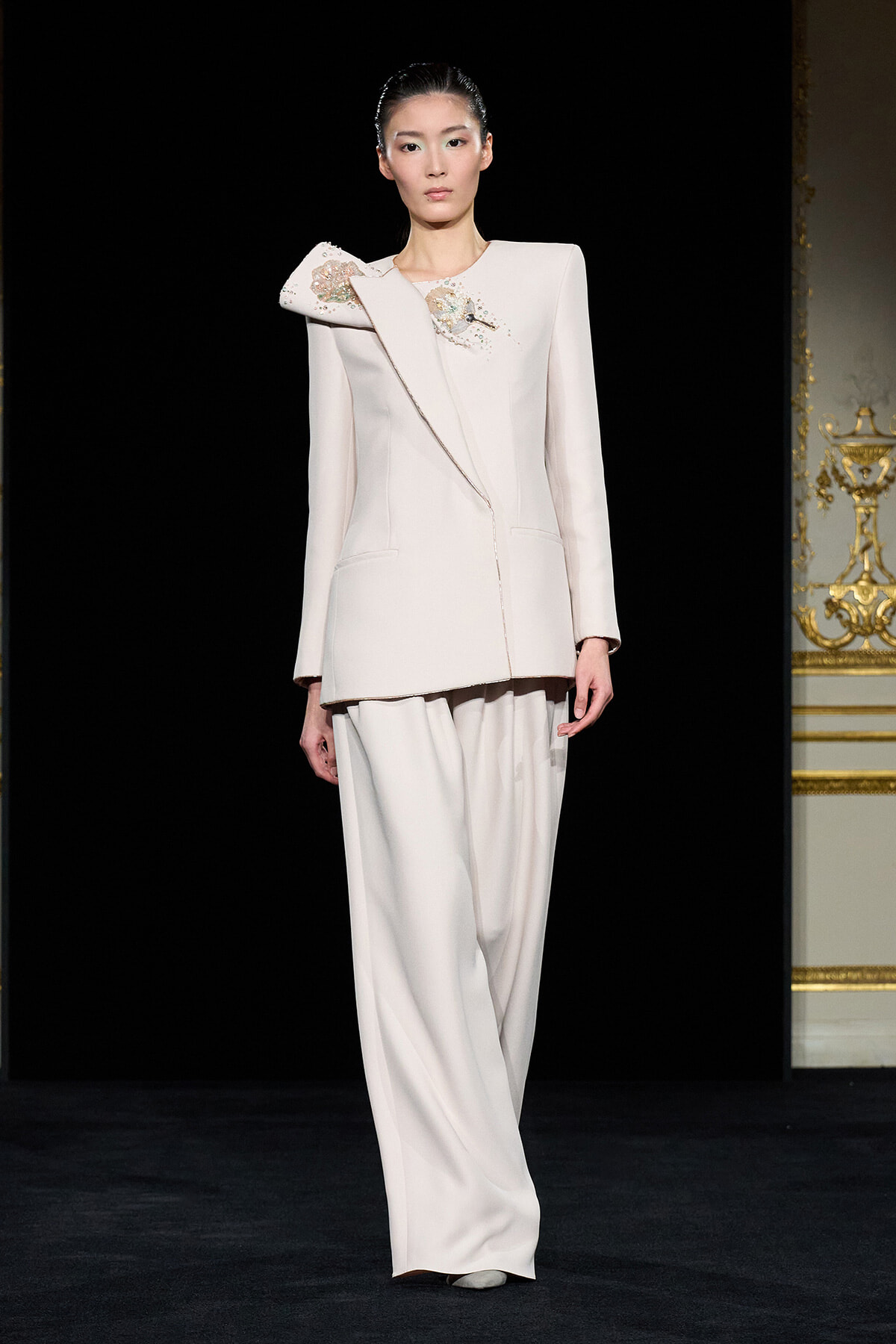 Model in a pale pink tailored blazer with a sculpted embroidered shoulder and matching wide-leg trousers on a runway surface.