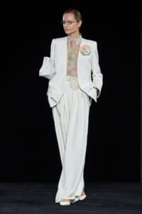 Model on a runway wearing a white suit with wide-leg trousers, sheer blouse, and pastel pocket square.",