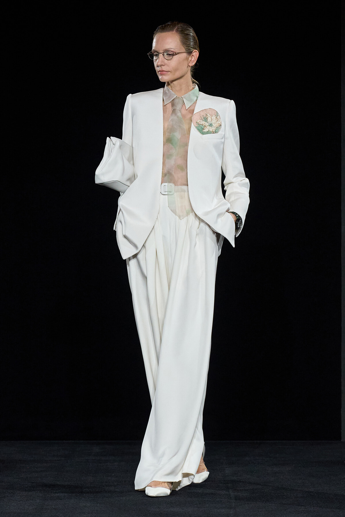 Model on a runway wearing a white suit with wide-leg trousers, sheer blouse, and pastel pocket square.",