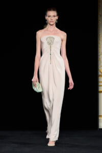 Model walks the runway in a strapless beige gown with a jeweled belt-like embellishment at the bust and a draped skirt, clutching a mint-green bag.