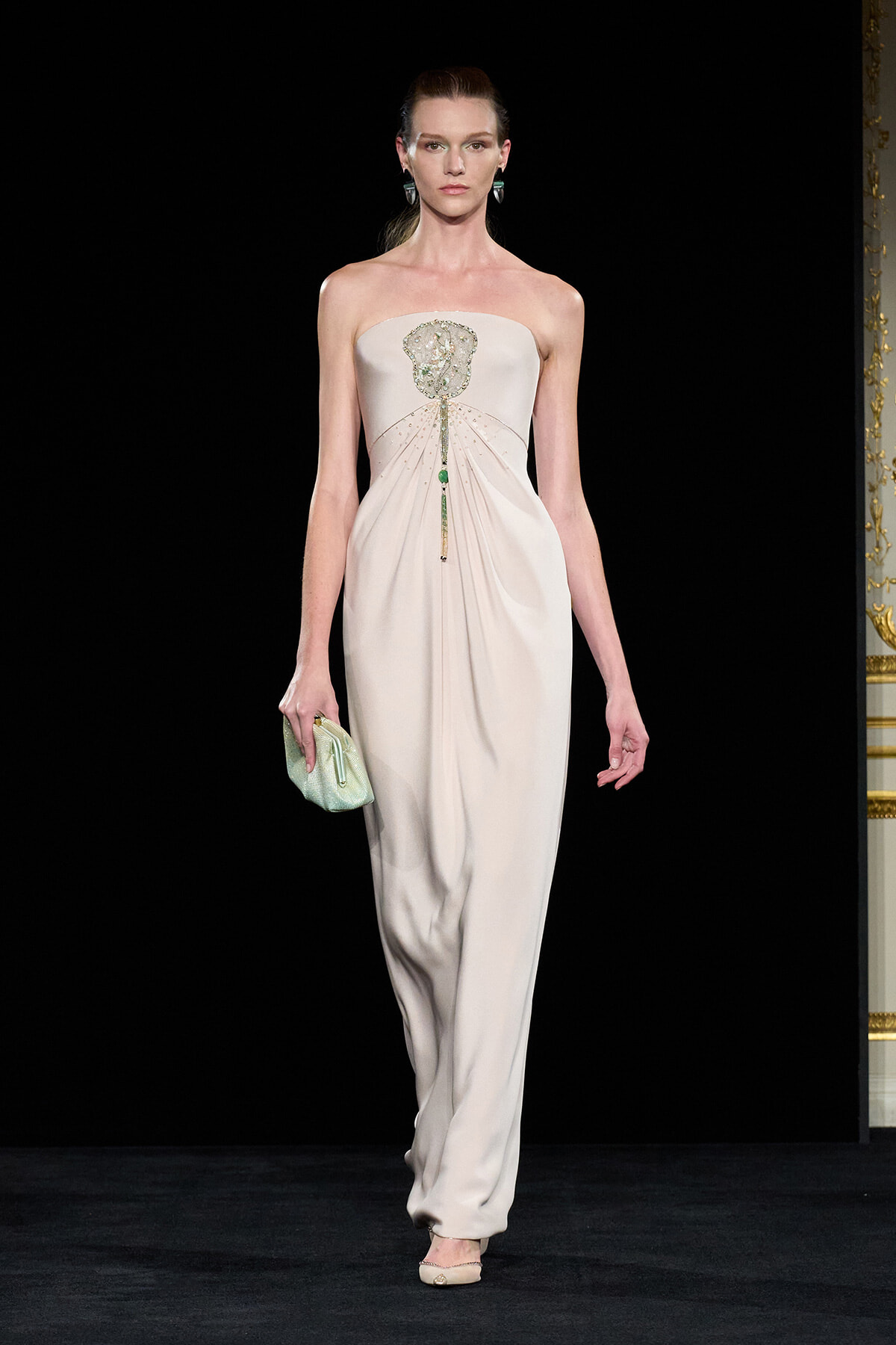 Model walks the runway in a strapless beige gown with a jeweled belt-like embellishment at the bust and a draped skirt, clutching a mint-green bag.