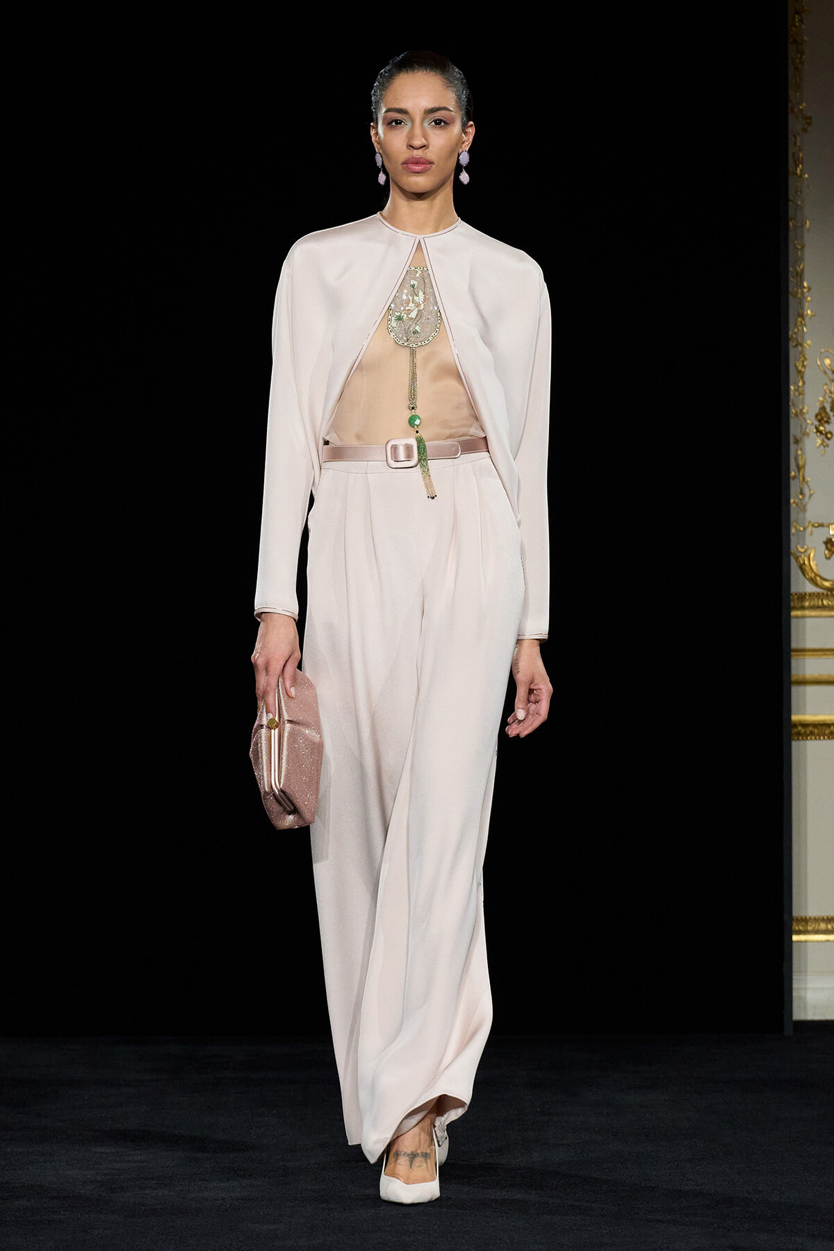 Model on a runway wearing a pale pink cape-front jumpsuit with a sheer top, embellished necklace, and matching pink clutch.