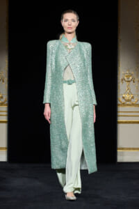 Runway model wearing a shimmering mint-green sequined long coat over pale green suit pants, on a fashion show runway.