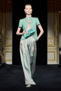 Model walking a runway in a mint-green beaded top, gradient satin wide-leg pants, teal clutch, and matching green jewelry.
