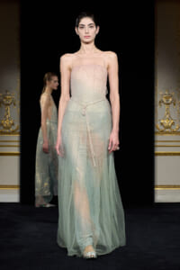 Model on a runway wearing a pale mint, beaded strapless gown with a sheer overlay and a thin belt, ornate gold-trimmed backdrop behind.