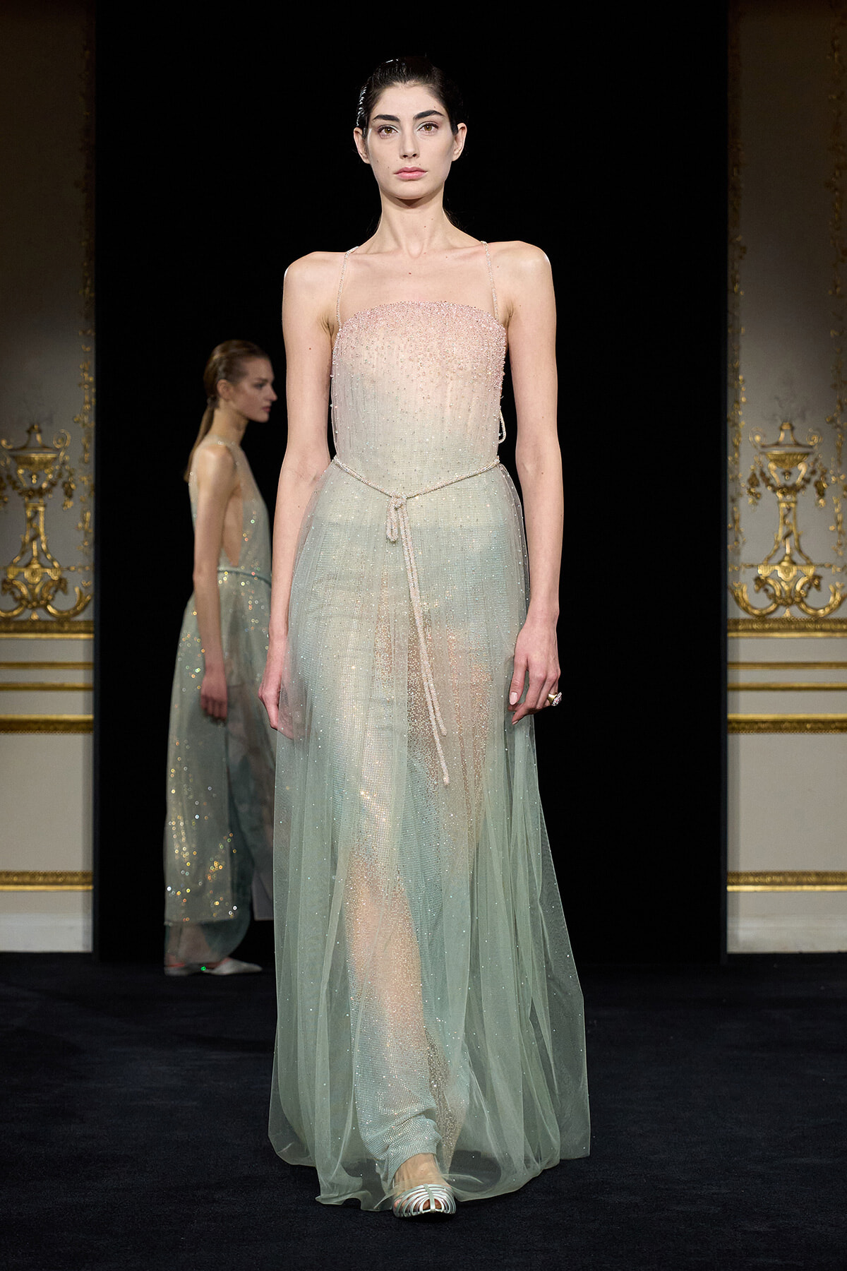 Model on a runway wearing a pale mint, beaded strapless gown with a sheer overlay and a thin belt, ornate gold-trimmed backdrop behind.