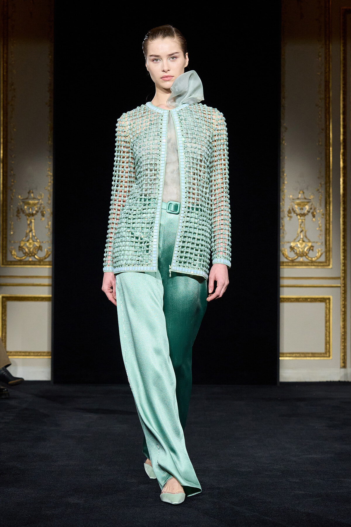 Model walking a runway in a mint-green beaded cardigan over a sheer top with satin wide-leg pants.