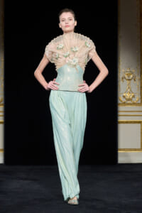 Model on runway wearing a pale mint-green glitter jumpsuit with a sculpted sheer ruffled capelet embroidered with floral appliques.