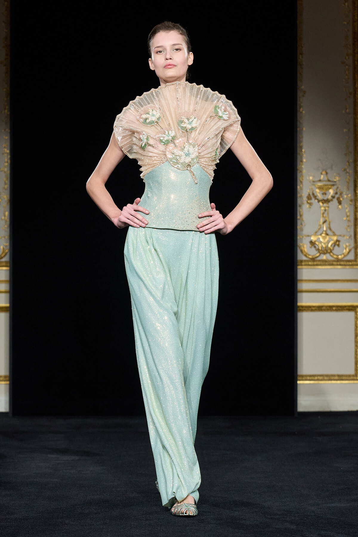 Model on runway wearing a pale mint-green glitter jumpsuit with a sculpted sheer ruffled capelet embroidered with floral appliques.