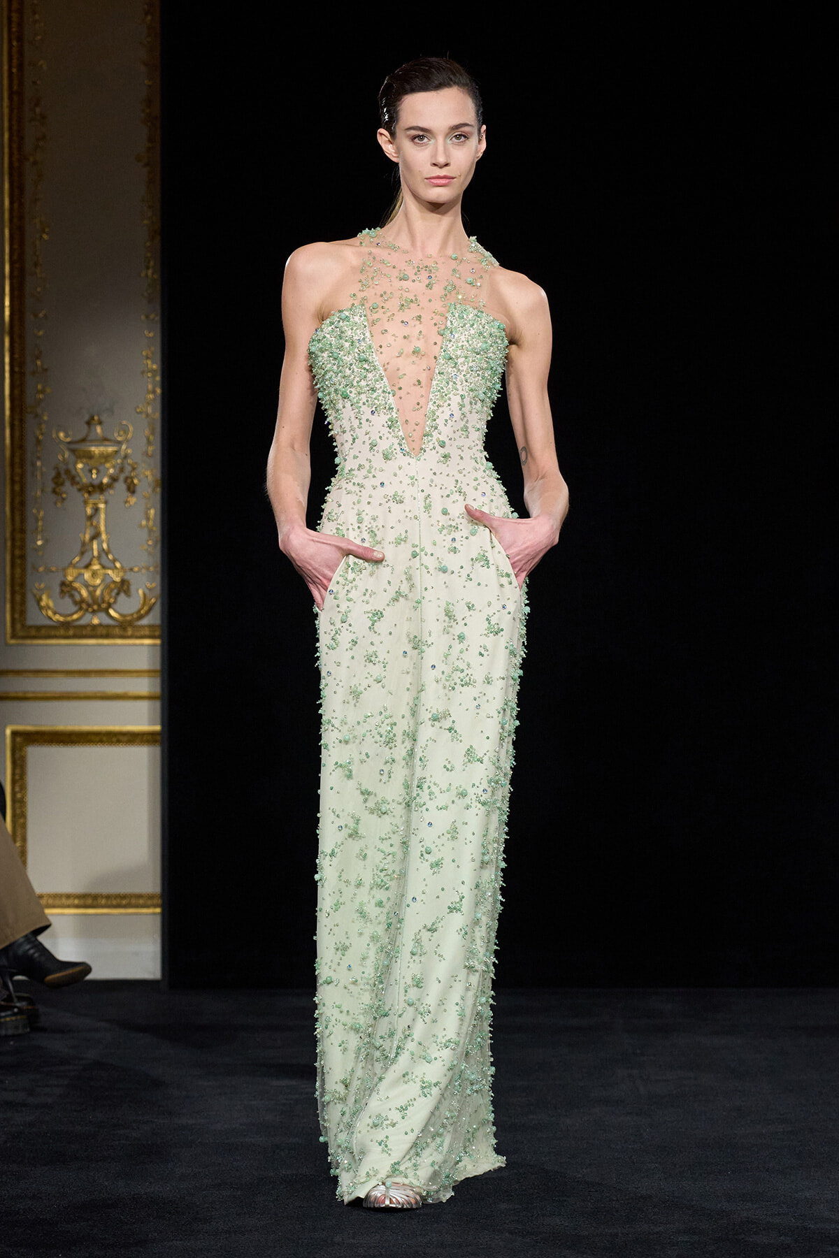 Model walking the runway in a mint-green, beaded gown with a sheer plunging bodice and pockets at the hips.