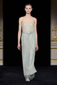 Model walks a shimmering pale blue, floor-length gown with spaghetti straps and a matching belt on a runway.