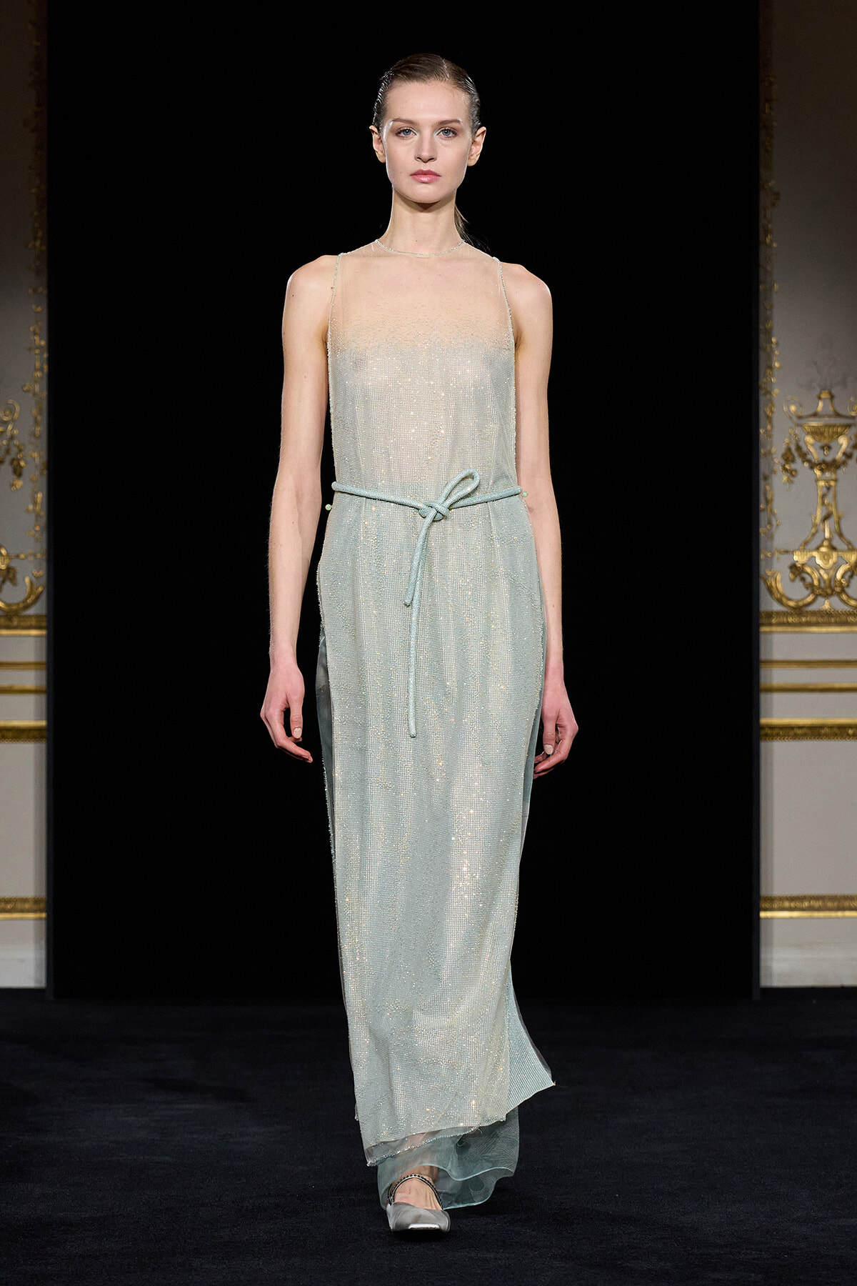 Model walks a shimmering pale blue, floor-length gown with spaghetti straps and a matching belt on a runway.
