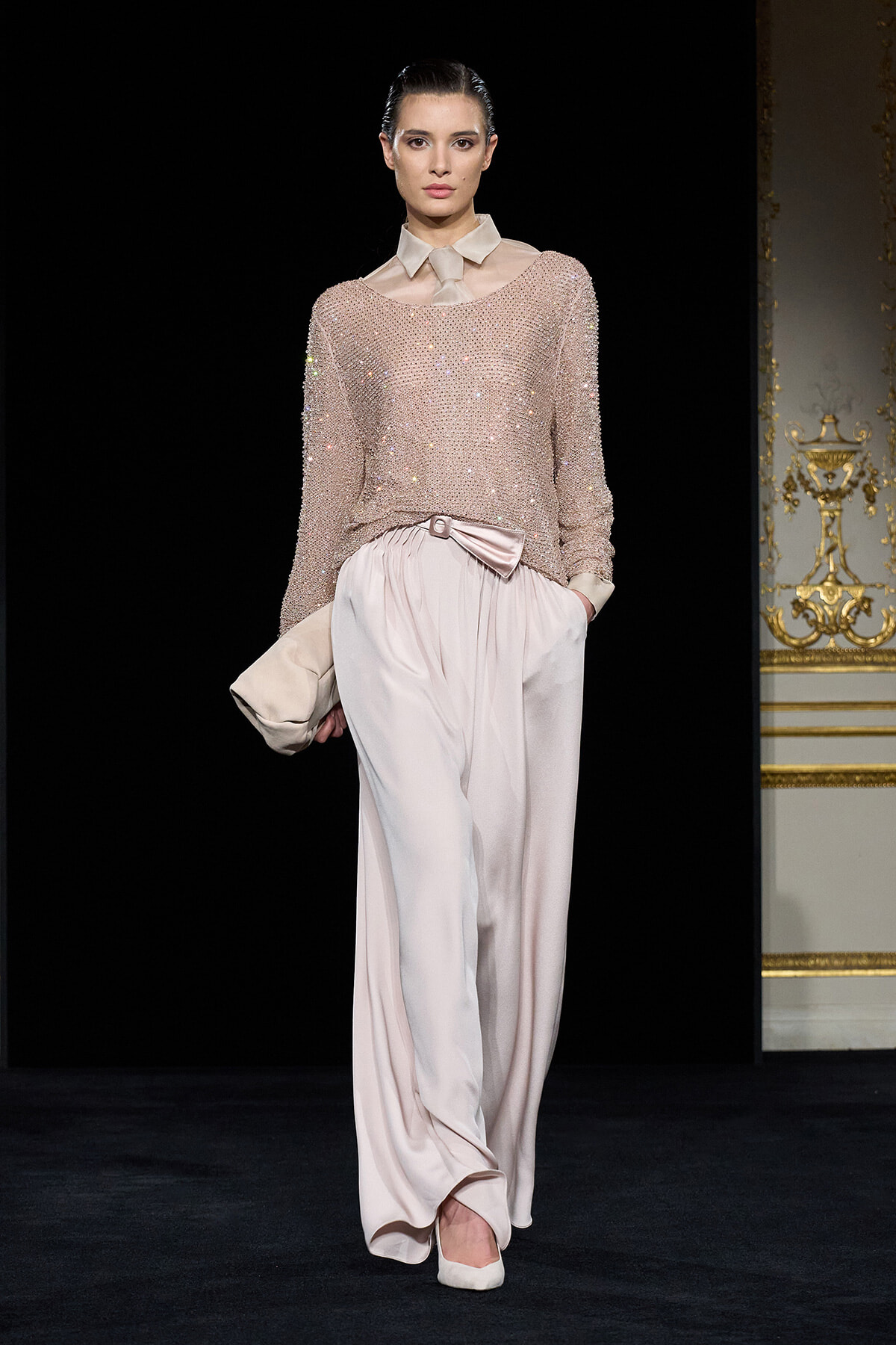 Model walking a runway in a pale pink sequined knit top with a cream collar, paired with flowing satin pink wide-leg pants and a bow belt.