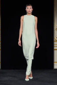 Model walks a runway in a pale mint-green sleeveless gown with beaded side panels and matching heels against a black backdrop.
