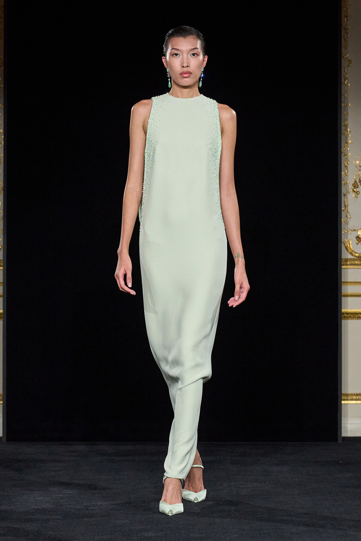 Model walks a runway in a pale mint-green sleeveless gown with beaded side panels and matching heels against a black backdrop.
