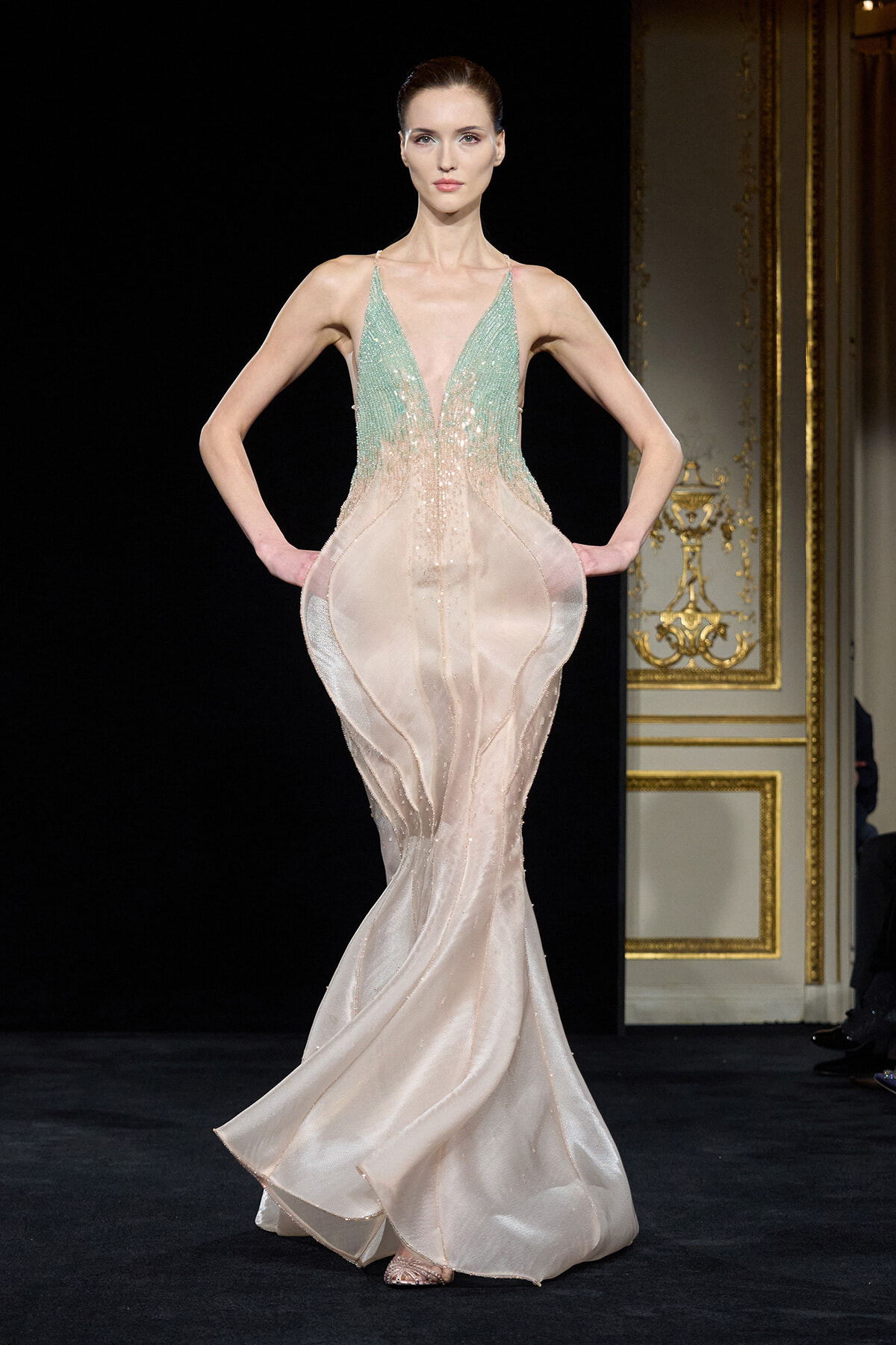 Model wearing a mint green, bead-embellished V-neck bodice gown that fades to pale pink, with a mermaid silhouette on a runway.