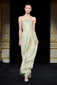 Model walking the runway in a strapless pale green beaded gown with a structured bodice and flowing, draped skirt against a black backdrop with ornate gold walls.