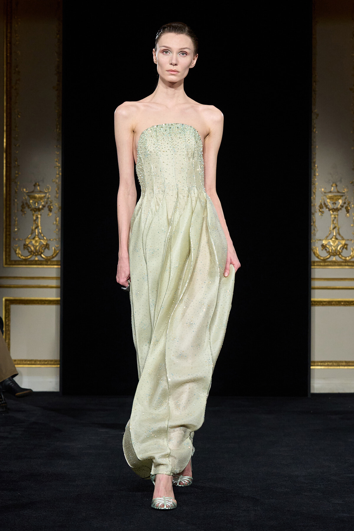 Model walking the runway in a strapless pale green beaded gown with a structured bodice and flowing, draped skirt against a black backdrop with ornate gold walls.