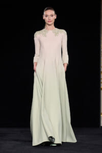 Model on runway wearing a pale, long-sleeved gown with green leaf embroidery at the shoulders and a flowing, floor-length skirt.