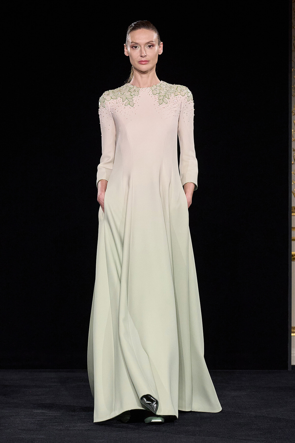 Model on runway wearing a pale, long-sleeved gown with green leaf embroidery at the shoulders and a flowing, floor-length skirt.