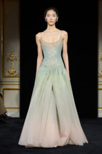 Model on a runway wearing a gradient aqua to pale pink beaded gown with thin straps and emerald earrings on the catwalk.