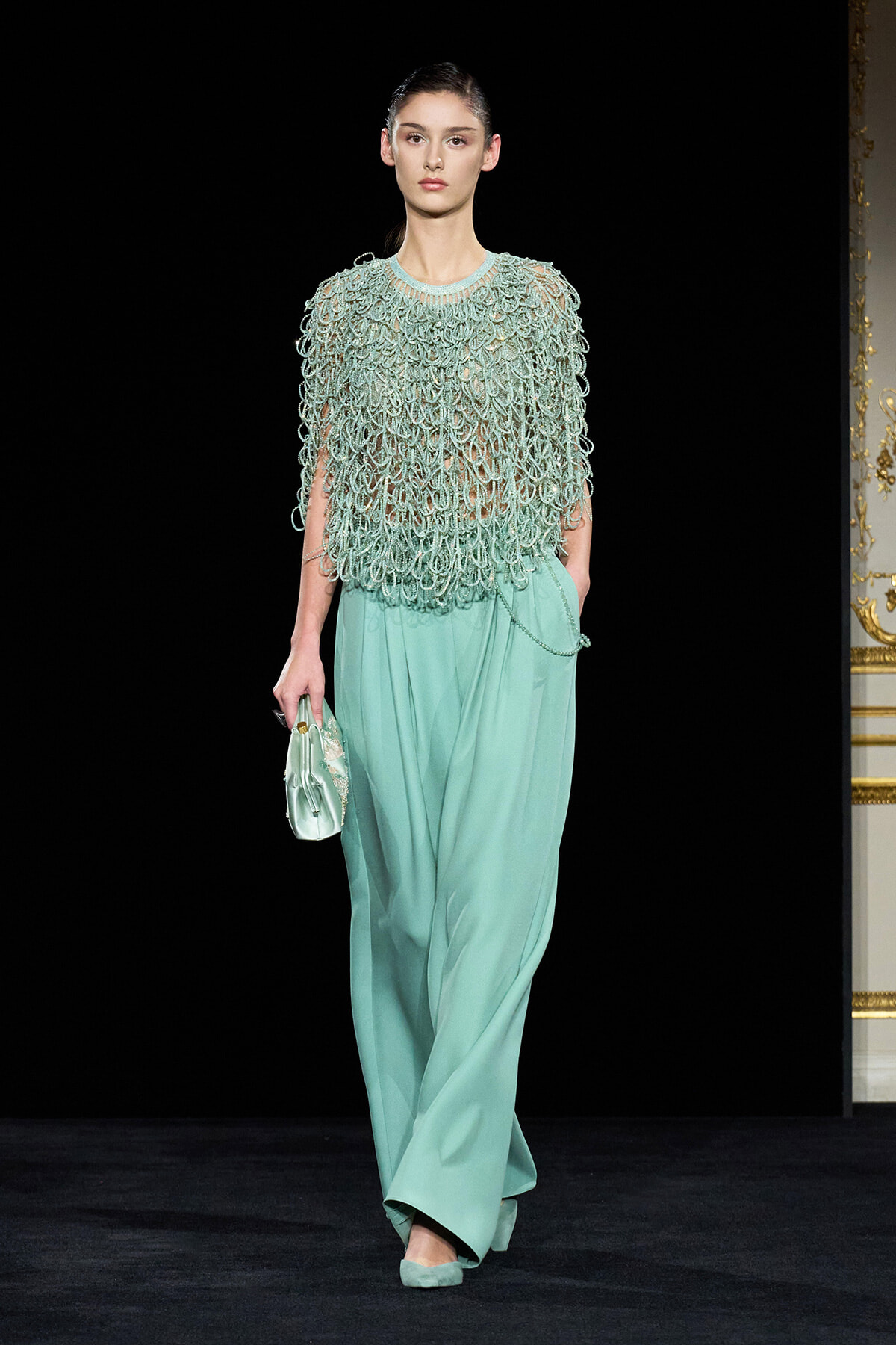 Model walking runway in a mint-green ensemble: oversized fringe-knit top with loop details over wide satin trousers, matching clutch and shoes.